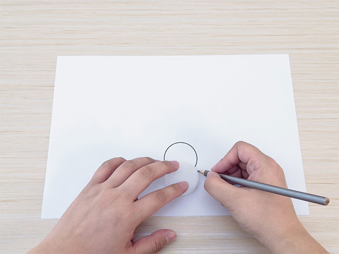Two hands are shown tracing a circle on a white sheet of paper using a gray pencil. The left hand holds a white bottle cap against the paper, using it as a template, while the right hand guides the pencil around the edge of the cap. The paper rests on a light wood-grain surface. This close-up image suggests a simple crafting or drawing activity.