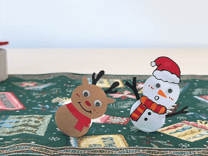 A small paper reindeer and a paper snowman wobble gently on a green patterned surface. The reindeer is brown with black antlers, googly eyes, a red nose, and a red scarf. The snowman is white with black twig arms, googly eyes, an orange carrot nose, a red and orange striped scarf, and a red Santa hat. The background suggests a festive, possibly Christmas-themed setting.