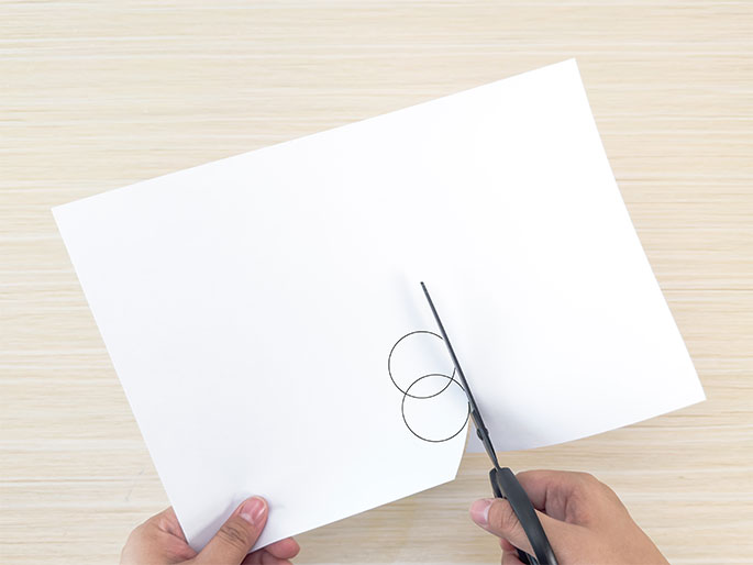 A pair of hands is shown cutting a white sheet of paper with black scissors. The left hand holds the paper, which rests on a light wood-grain surface, while the right hand operates the scissors. Two pencil-drawn circles are visible on the paper, and the scissors are cutting along the edge of one of them. This image depicts the process of cutting out a circular shape from paper.