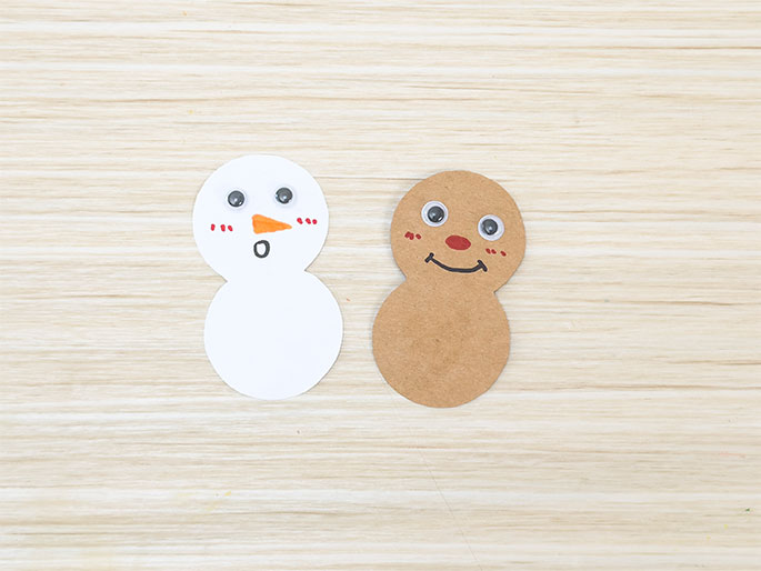 light wood-grain surface. On the left, a white cutout resembles a snowman, with a small orange triangle for a nose, rosy cheeks, and a small o for a mouth. On the right, a light brown cutout, possibly intended as a gingerbread person, has a red circular nose, rosy cheeks, and a smiling mouth. These figures appear to be part of a childrens craft project.