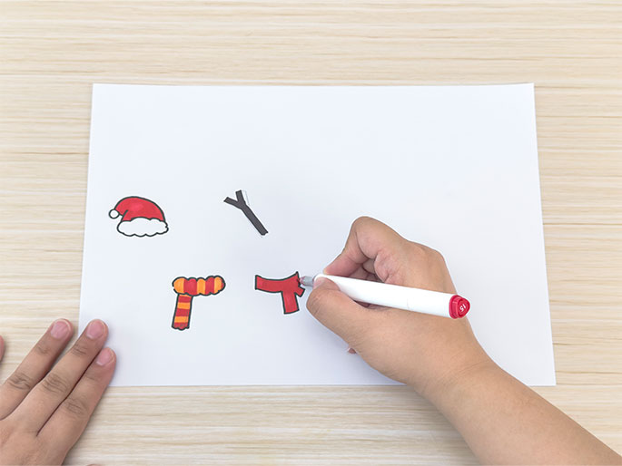 On a piece of white paper lying on a light wooden table, a hand holding a red marker is coloring in a drawing of a red scarf. Other simple drawings, including a Santa hat, a stick arm, and an orange and yellow striped scarf, are also visible on the paper. Another hand is partially visible on the left side of the frame. The scene suggests a crafting or drawing activity, likely related to creating snowman decorations.