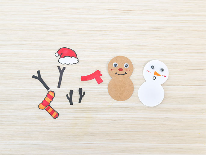 Arranged on a light wood-grain surface are various paper cutouts, seemingly for a snowman and gingerbread person craft. A white snowman figure with googly eyes, an orange paper nose, and drawn rosy cheeks and mouth is positioned on the right. Next to it is a light brown gingerbread figure, also with googly eyes, a red paper nose, and drawn features. Scattered above and to the left are additional cutouts: a red and white Santa hat, two black stick arms, a red paper scarf, a striped orange and yellow paper scarf, and two black mitten shapes. The arrangement suggests the components of a children's craft project, ready to be assembled.