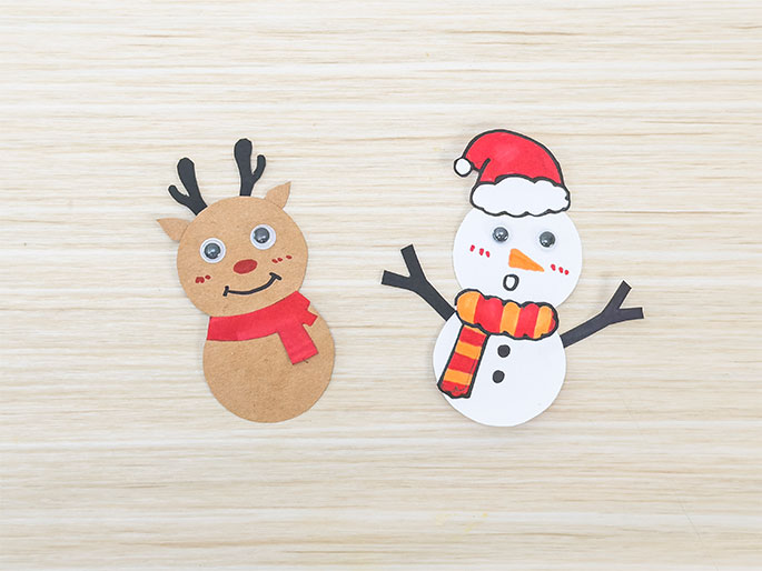 Two completed holiday-themed crafts rest on a light wood-grain background. On the left, a reindeer figure made from light brown card stock features googly eyes, a drawn red nose and smiling mouth, black card stock antlers, and a red paper scarf. On the right, a snowman crafted from white card stock includes googly eyes, a drawn orange carrot nose, rosy cheeks, and an o shaped mouth. The snowman also wears a striped orange and yellow paper scarf, a red paper Santa hat, and has black card stock arms. These simple, child-made decorations evoke a festive spirit.