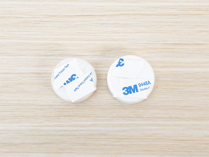 Two white adhesive discs are placed on a light-colored wood-grain surface. The disc on the right has a partially peeled backing, revealing the 3M logo and product number 9448A, indicating it's 3M double-sided tape. The disc on the left has a different backing, partially peeled to show blue text indicating coated tissue tape. These discs are ready to be used for mounting or sticking items.