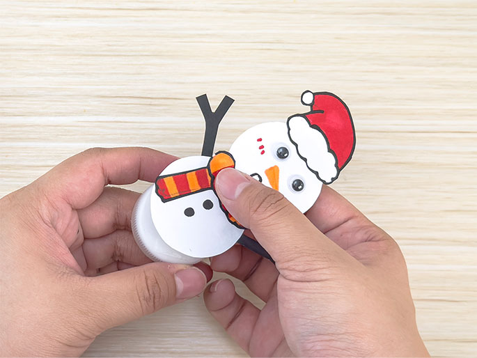 Two hands are shown assembling a snowman craft against a light wood background. The snowman's body, a white circle with drawn features (googly eyes, an orange carrot nose, rosy cheeks, black buttons, and a surprised expression), is being attached to a white plastic bottle cap. The snowman wears a red and white Santa hat and a striped orange and yellow scarf. One hand holds the bottle cap while the other hand positions the snowman cutout, suggesting the process of gluing or taping the pieces together. Black stick arms are also attached to the snowman figure.
