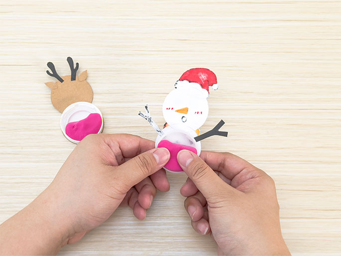 Hands are shown in the process of making Christmas crafts on a light wooden surface. In the center, a snowman figure made from a white circle with drawn features (googly eyes, an orange carrot nose, rosy cheeks) and a red and white Santa hat is being attached to a white plastic bottle cap filled with pink putty or clay. Black stick arms are affixed to the snowman. To the left, another bottle cap with pink putty is visible, with a partially completed reindeer head (light brown card stock with black antlers) positioned above it. The scene captures the midway point of creating these festive decorations.