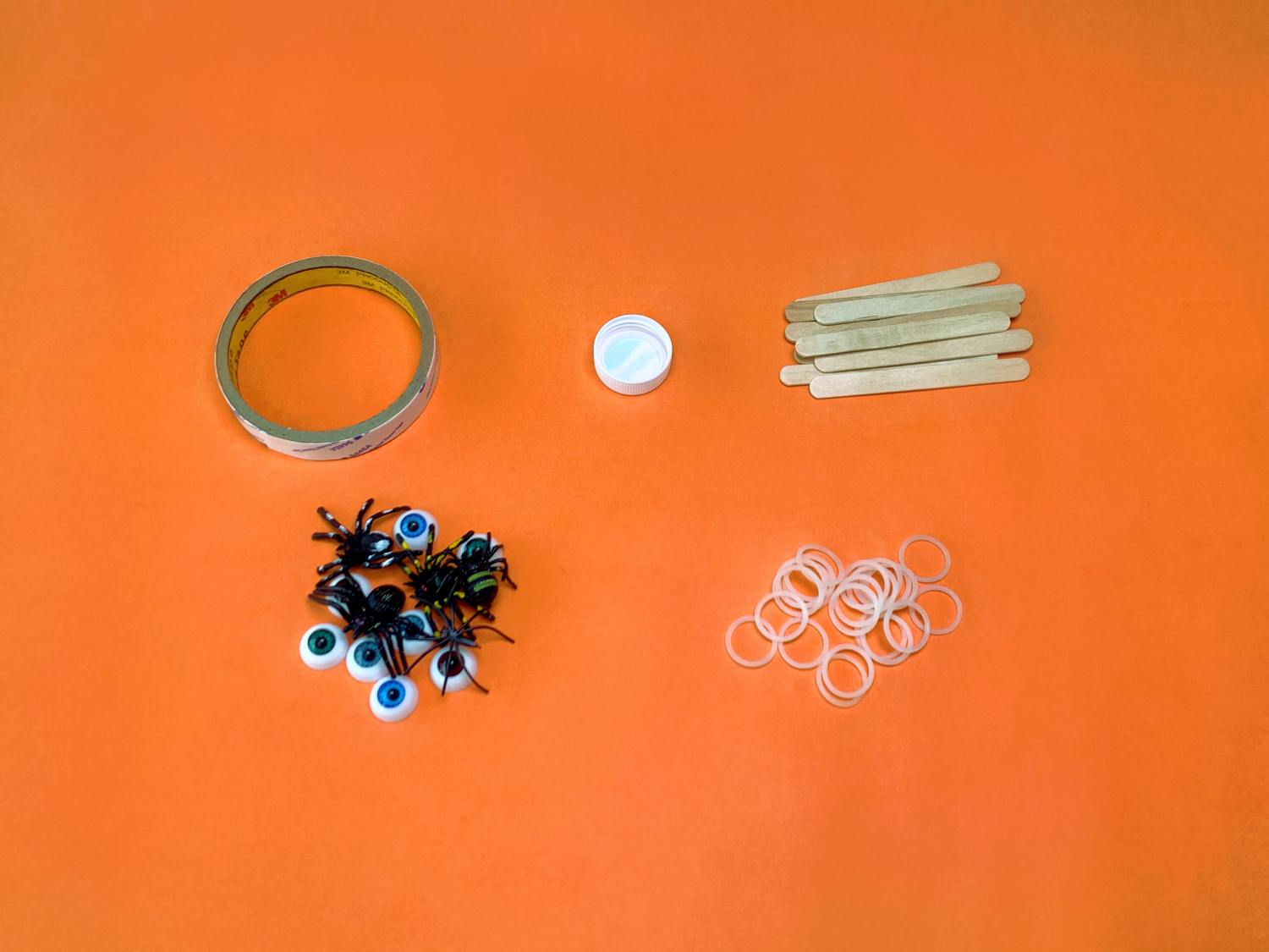 Gather your spooky supplies! A roll of tape, a bottle lid, craft sticks, elastic bands, plastic spiders and googly eyes are laid out on a bright orange surface, ready for catapult construction. Let's get building!