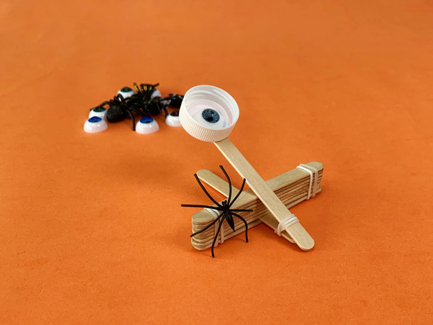 Prepare for a spidery launch! A completed spider catapult, made from craft sticks and a bottle top with a googly eye, is ready for action on a bright orange background, with extra spiders and googly eyes nearby. Let's get those creepy crawlies flying!