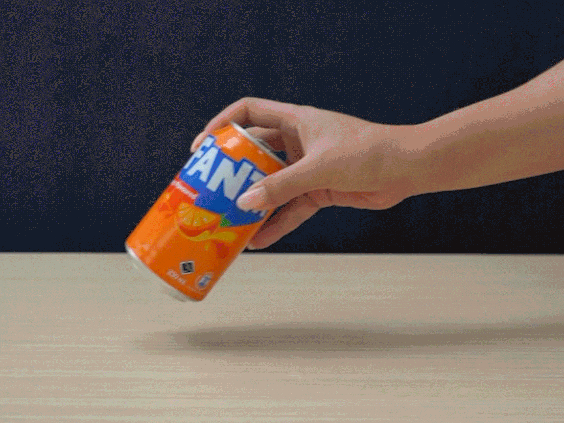A hand sets an orange Fanta can spinning on a light-coloured table against a dark blue background.