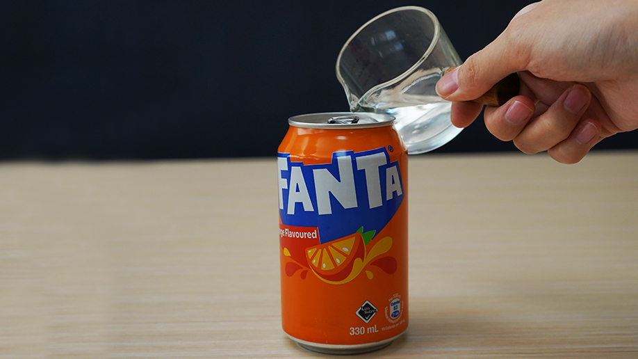 A hand pours water from a small, clear glass pitcher or jug into an open can of Fanta.