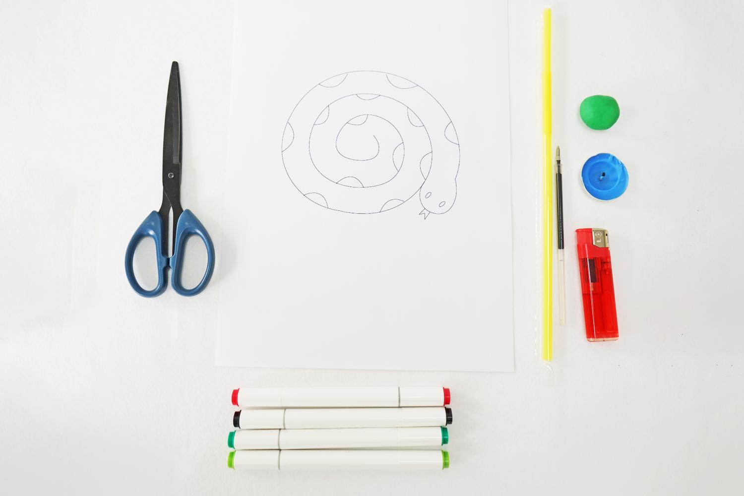 Everything's ready for a slithery science experiment: scissors, a snake template, markers, a straw, modelling clay, a small blue disc, a pen, and a lighter are neatly arranged. Let's get creative!