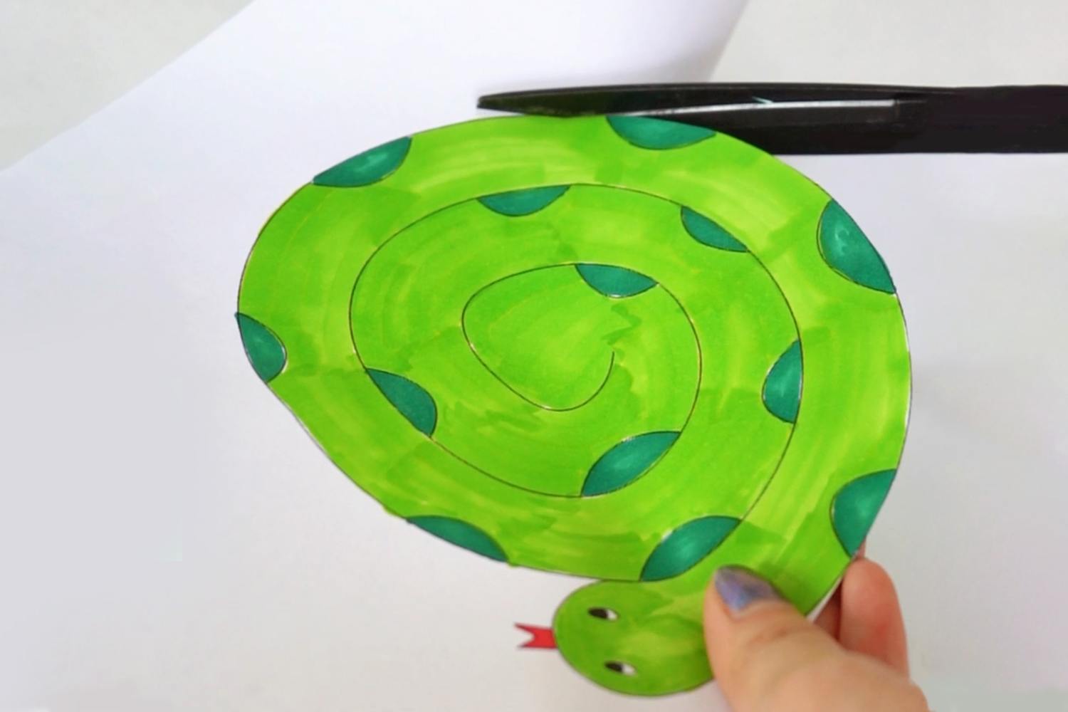 A hand holds a bright green paper snake, coiled and decorated with teal spots, recently cut out from a sheet of paper.