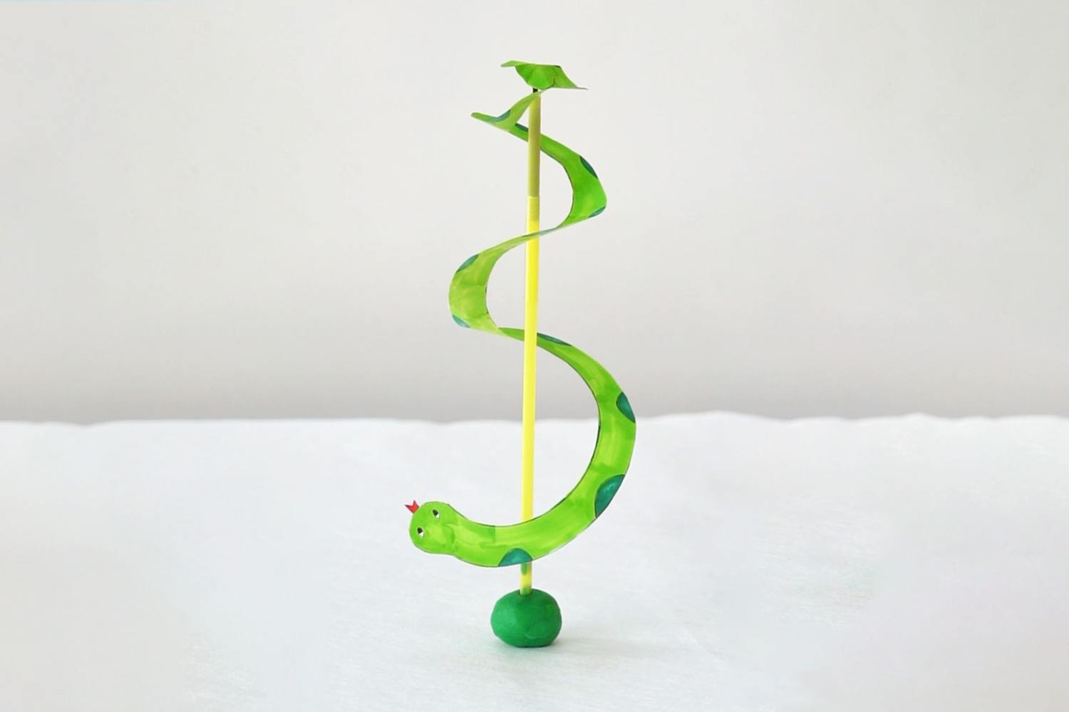 A whimsical, handcrafted paper snake, painted bright green with teal spots, spirals around a yellow straw, balanced on a small green clay ball; a playful and charming creation.