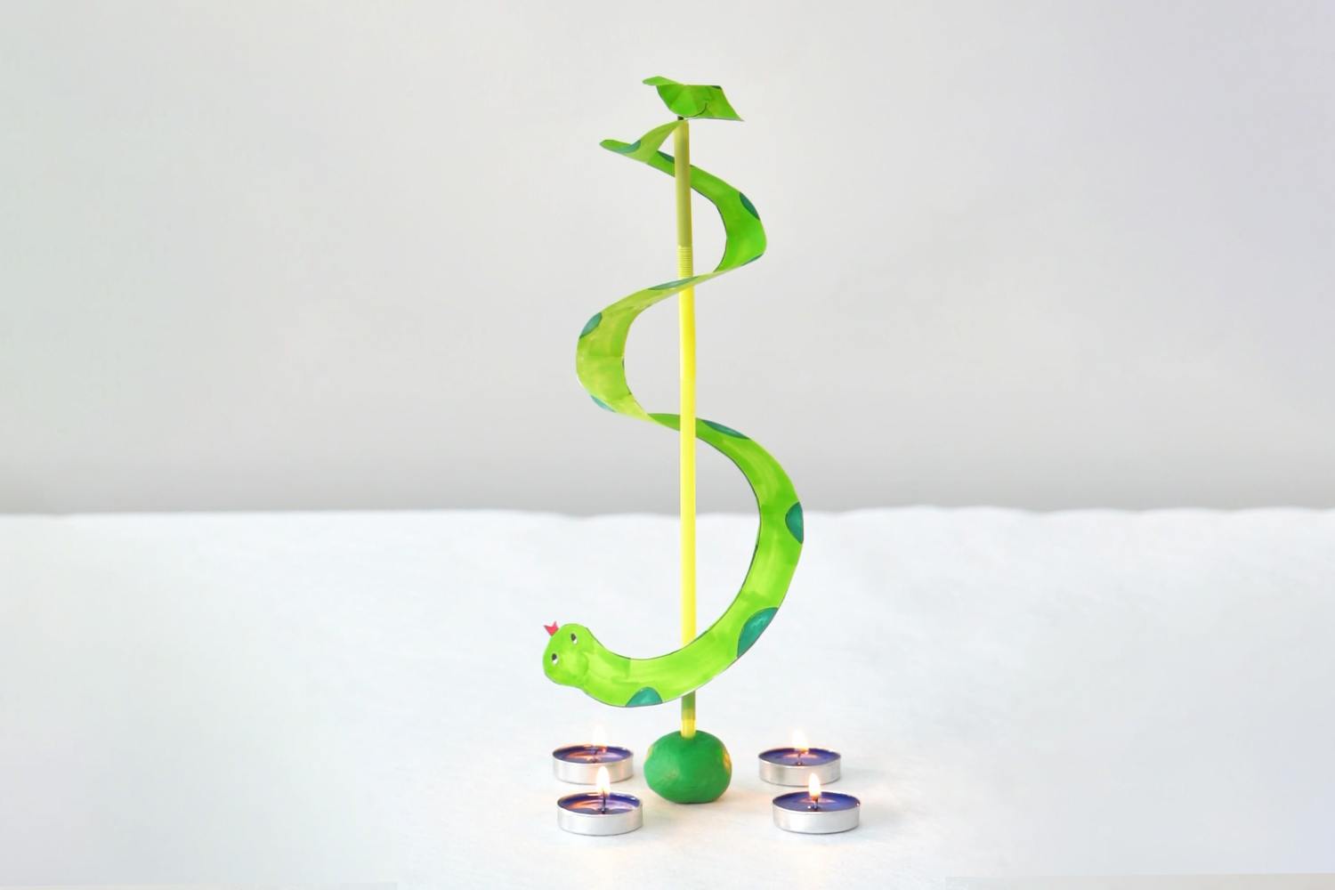 A playful, handcrafted paper snake, vibrant green with teal spots, spirals around a yellow straw, balanced delicately on a small green clay ball, encircled by four softly glowing teal candles on a white surface.