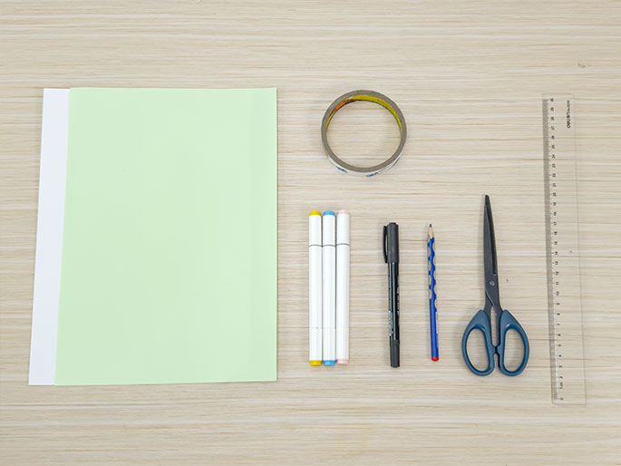 A flat lay arrangement of craft supplies on a light wood grain surface. The supplies include a sheet of light green paper, a roll of clear tape, three white markers with colored caps (light blue, pink, and yellow), a black pen, a blue pencil, a pair of scissors, and a clear plastic ruler. These items suggest preparation for a paper craft or similar activity.