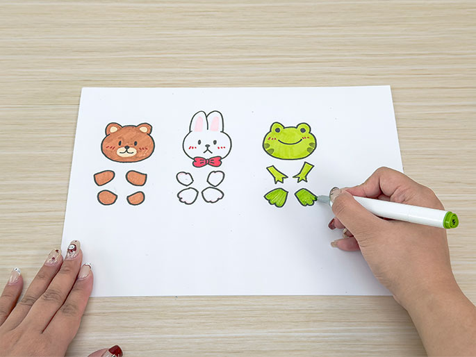 On a piece of white paper against a light wooden background, simple, cartoon-like drawings of a bear, a rabbit, and a frog are being colored in. Each drawing is separated into parts – the head and limbs – as if to be cut out and assembled. A hand holding a light green marker is coloring in one of the frog's feet. The scene suggests a children's craft activity.