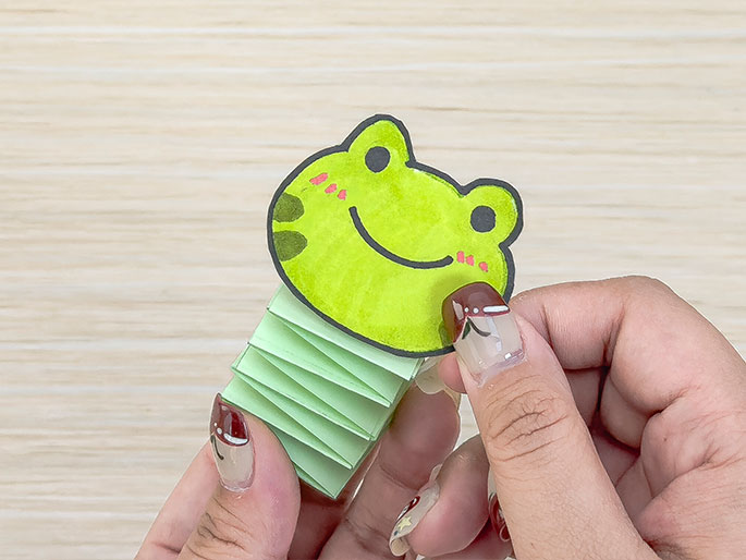 Hands hold a small paper craft of a frog. The frog's head is drawn and colored on light green paper, while its body is a folded, accordion-style stack of pale green paper. The light wood background suggests a children's craft or origami project.