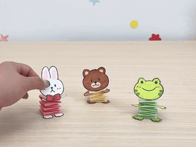 Three small, handmade paper animal figures stand on a light wood surface. A hand reaches out to touch the first figure, a white rabbit with a red bow tie. The second figure is a brown bear, and the third is a green frog. Each animal's body is made from a folded, accordion-style stack of paper. The image evokes children's crafts or simple origami.