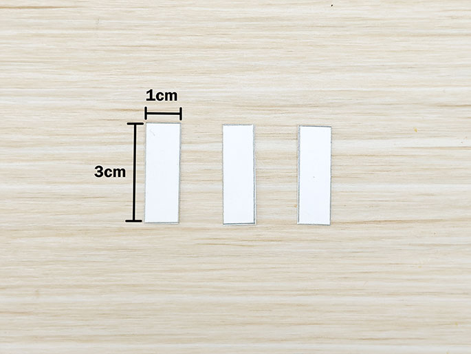 Three small, identical white rectangular strips of paper lie on a light-colored wooden surface. Lines and text indicate their dimensions: 3cm long and 1cm wide. The image suggests they are prepared for a craft project.