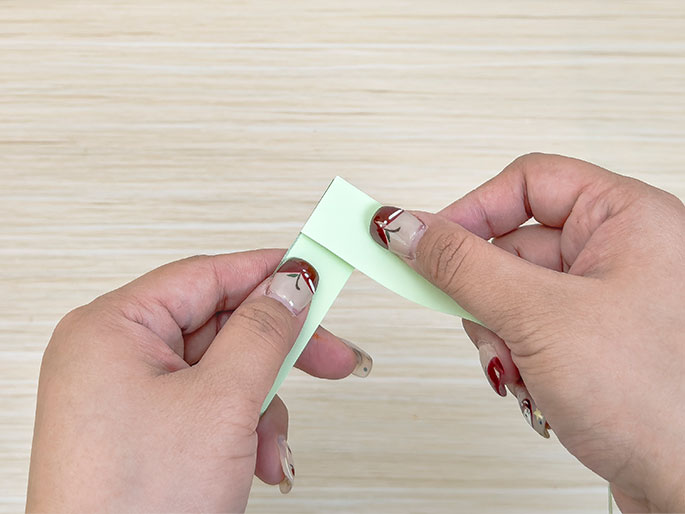 Two hands are folding a pale green strip of paper against a light wood background. The fingers hold the paper delicately, creating a bend, likely as part of a paper craft or origami project.