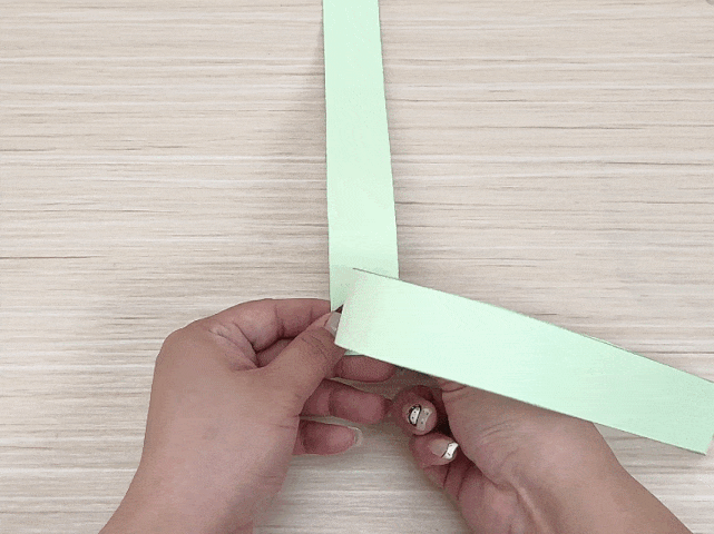 Two hands manipulate two pale green strips of paper on a light wood background. One strip is vertical, and the other is being woven horizontally around it, suggesting the creation of a simple paper weave or craft project.