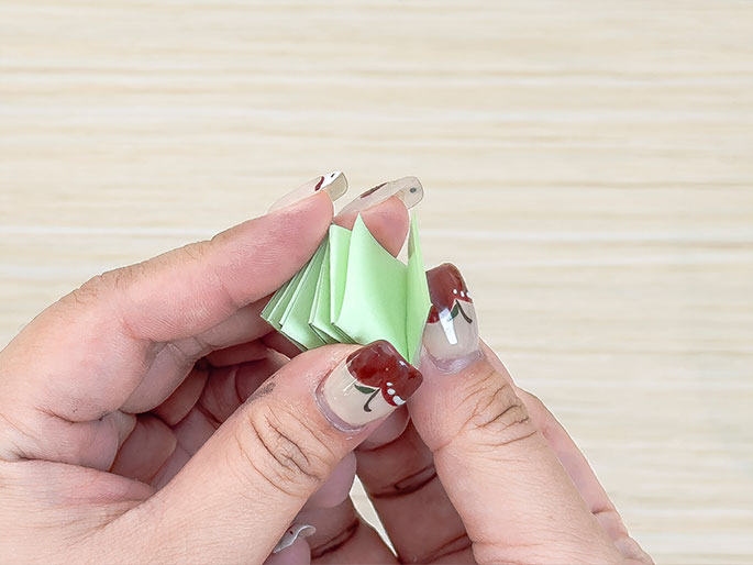 Hands hold a small, folded, light green paper object resembling a fan or flower. A clear plastic piece is attached at the top, likely securing the folds. The background is a light wood surface. The image suggests a stage in a paper craft project.