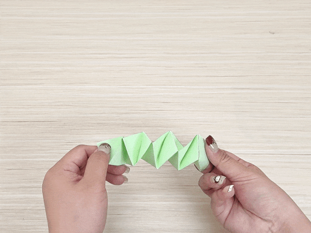 Two hands hold a light green paper creation resembling a folded, zig-zagging ribbon or caterpillar. The paper is folded into multiple triangular segments. The background is a light wood surface, suggesting this is a paper craft project.