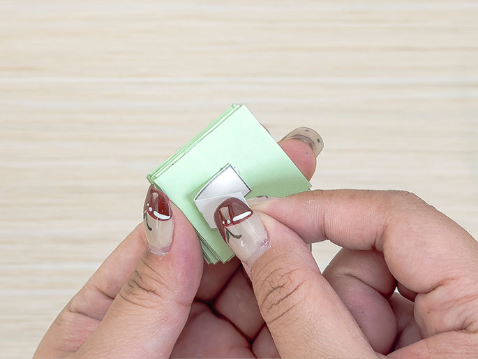 Two hands hold a stack of folded, pale green paper squares. A piece of double-sided tape is being applied to one of the folded sections. The light wood background suggests a crafting or DIY project in progress.