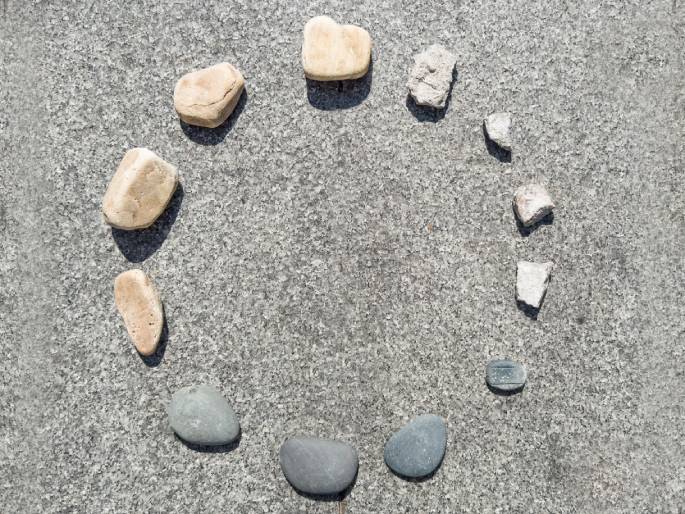Various rocks and pieces of broken concrete are arranged in a circle on a textured, gray surface, likely concrete or asphalt. Smooth, light-brown rocks alternate with smaller, jagged pieces of light gray concrete. Several darker gray, smooth stones are grouped together on the lower part of the circle. The arrangement suggests the face of a clock or a marker for a game.