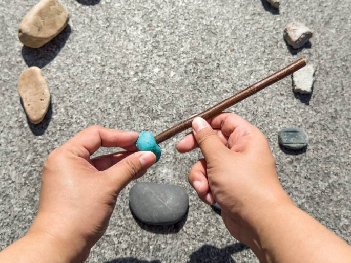 Two hands hold a thin, brown stick with a small ball of turquoise-colored putty or clay affixed to one end. The background is a textured gray surface, likely concrete or asphalt, and several rocks and pieces of broken concrete are scattered around, out of focus. The hands appear to be in the process of attaching or securing the putty to the stick, possibly as part of a craft or game.