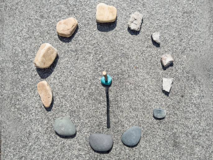 A homemade sundial is crafted on a gray, textured surface, likely concrete or asphalt. A central stick, anchored with a light blue base, casts a distinct shadow. Around the stick, a circle is formed by various rocks and broken pieces of concrete, serving as hour markers. The lighter-colored, smoother rocks are interspersed with the rougher, lighter gray pieces of concrete. The sundial appears to be indicating a time in the early afternoon.