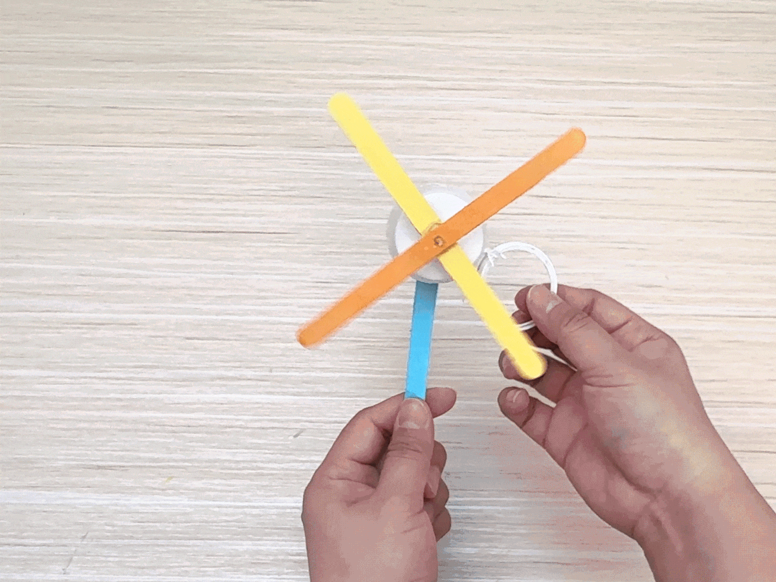 A hand holds a small, homemade toy resembling a whirligig or spinner. The toy appears to be constructed from a small, round object, possibly a bottle cap or button, attached to a light blue stick or handle. Yellow paper blades are affixed to the round object. A white string is tied to the handle and looped into a ring at the other end. The hand pulls the string back and forth, causing the yellow blades to spin rapidly, creating a blur of motion. The video demonstrates a simple DIY toy that can be made with readily available materials.