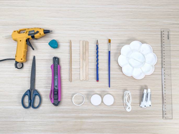 An assortment of craft supplies is neatly arranged on a light woodgrain surface. The items include a yellow hot glue gun, a small lump of teal putty or clay, a pair of blue scissors, a pink utility knife, thin wooden sticks, a pencil, a blue paintbrush, a white paint palette, a clear plastic ring, two small white discs, a length of white string or twine, two small tubes of glue, and a clear plastic ruler. These materials appear to be laid out in preparation for a craft project.