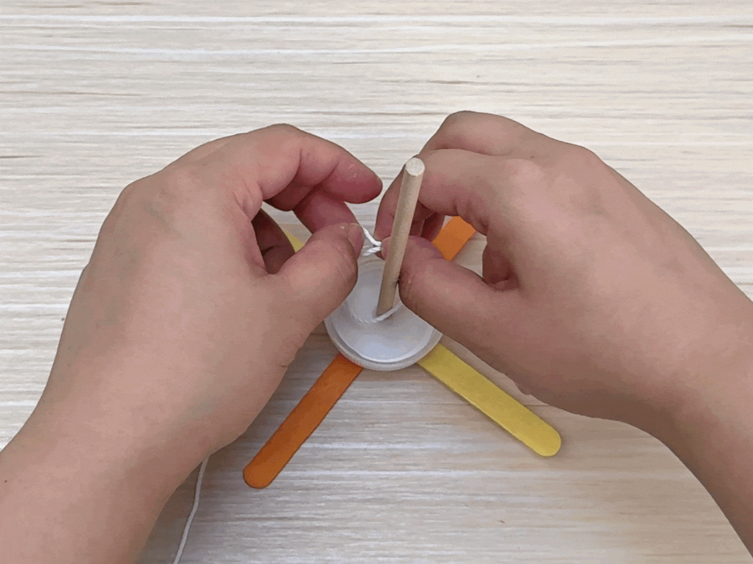 Two hands work on a craft project on a light wood grain surface. They are attaching a piece of string or twine to a small, central wooden dowel. The dowel is fixed to a white plastic lid or base, and several colorful craft sticks – orange and yellow – extend outwards from the base. The image captures the process of tying or securing the string, a key step in the construction of this DIY project.