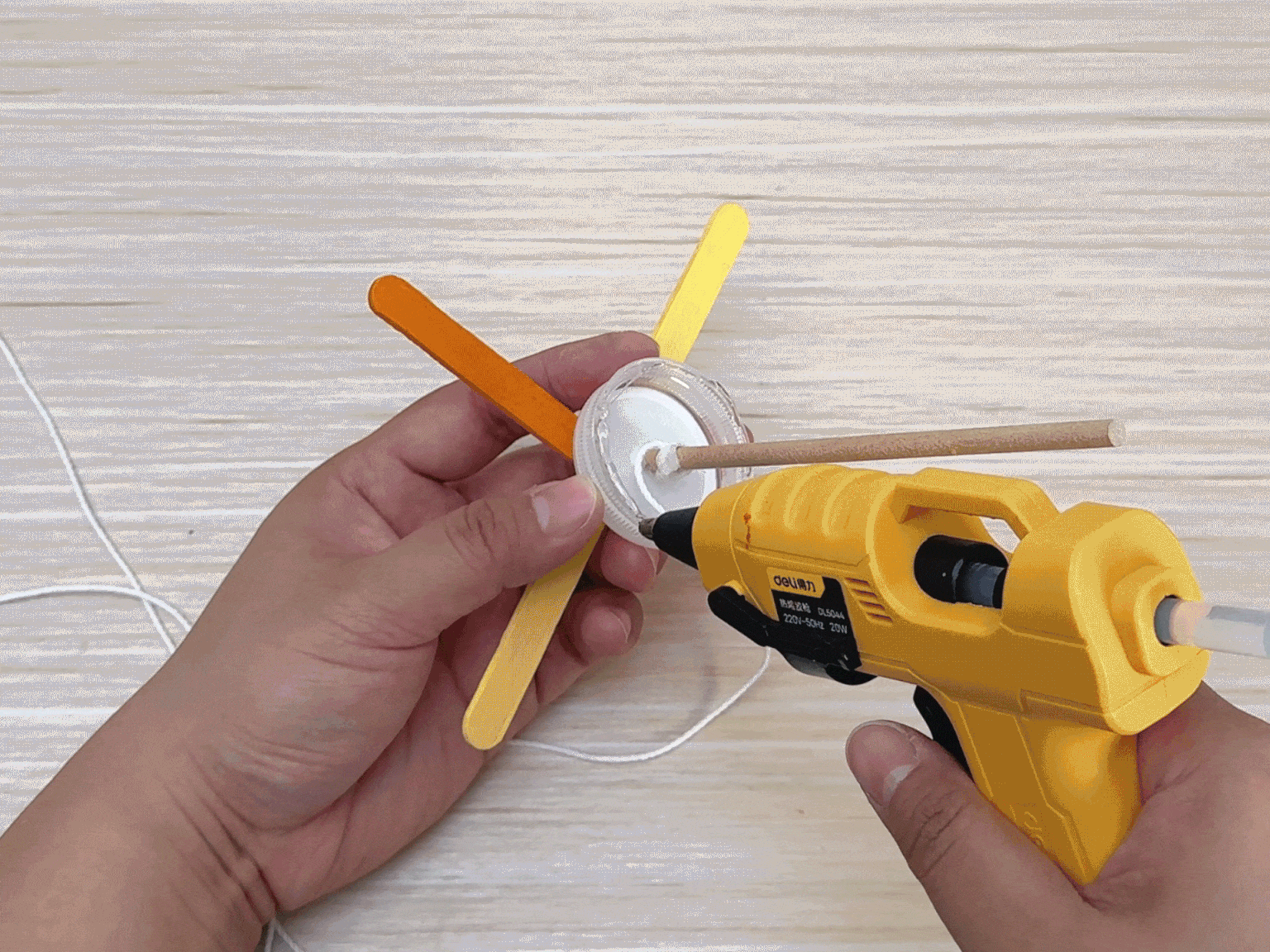 Two hands are constructing a craft on a light wood grain surface using a hot glue gun. One hand holds the craft, which consists of colorful craft sticks (orange and yellow) attached to a small white plastic lid or container. A wooden dowel extends from the center. The other hand operates a yellow hot glue gun, applying glue to the base of the dowel. A piece of string is also visible, likely part of the project. This image captures a mid-process step in creating the craft.