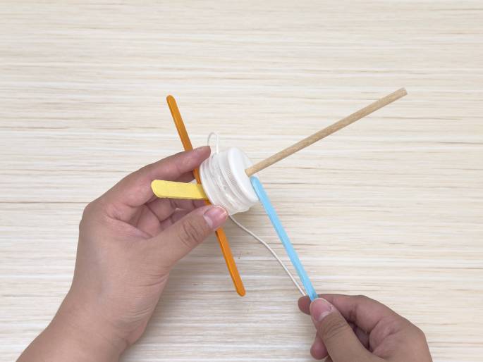 Two hands manipulate a partially completed craft project on a light wood grain surface. One hand holds the main body of the craft, which consists of a small white plastic container or lid with a wooden dowel inserted through it. Orange and yellow craft sticks are attached to the container. The other hand holds a light blue craft stick, seemingly preparing to attach it to the assembly. A piece of string or twine is wrapped around the container and extends outwards, suggesting it will play a role in the finished craft's function.