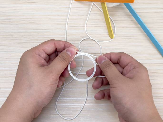 Two hands hold a length of white string or twine and a white plastic ring against a light wood grain background. The string is wrapped multiple times around the ring. In the background, partially out of focus, are colorful craft sticks (yellow and light blue), suggesting they are part of a larger craft project. The hands appear to be securing the string to the ring, indicating a step-by-step assembly process.