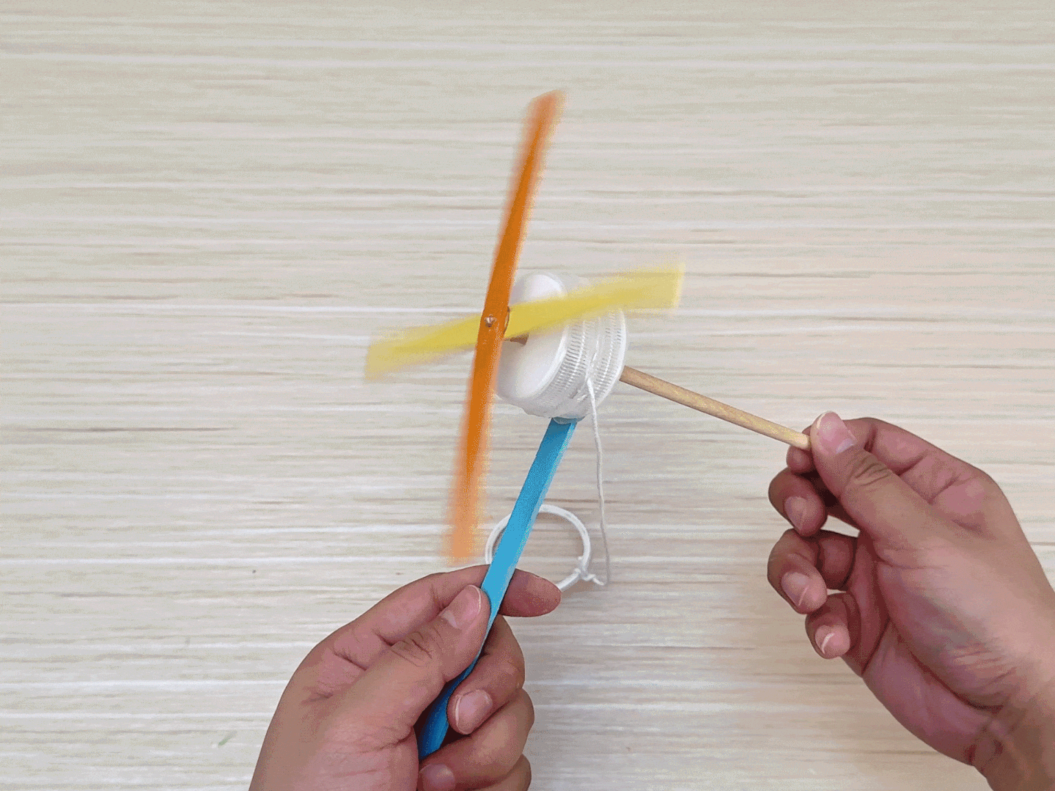 Two hands interact with a spinning, homemade toy against a light wood grain background. The toy resembles a small windmill or propeller. It features a central white plastic container or lid with crossed orange and yellow craft sticks forming the blades. A wooden dowel acts as the handle or axle. A light blue craft stick and a white string loop are also attached, suggesting they are part of the mechanism used to spin the toy. The blurred motion of the orange and yellow sticks indicates the toy is actively spinning.