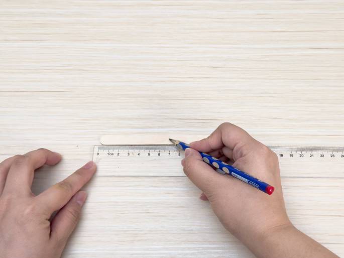 Two hands are shown on a light-colored wooden surface. A clear plastic ruler is positioned horizontally, and a small wooden craft stick lies on top of it. One hand holds the ruler in place at the zero mark, while the other hand uses a blue pencil to mark a line on the craft stick, guided by the ruler. The image appears to depict the first steps in a craft project, measuring and marking a wooden stick for cutting or other modification.