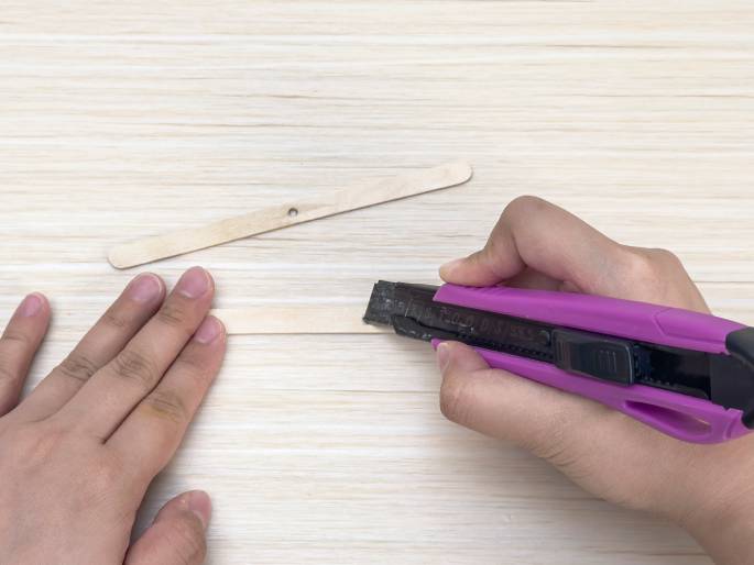 Two hands are working with small wooden craft sticks on a light woodgrain surface. One hand holds a craft stick flat against the surface, while the other hand uses a pink utility knife to carefully score or cut the stick. A previously cut stick with a small hole near one end lies above, suggesting a step in a craft project involving cutting and modifying these wooden pieces.