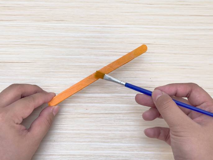 Two hands are painting an orange craft stick on a light wood grain surface. One hand holds the stick while the other hand applies orange paint with a small blue paintbrush. The focus is on the process of painting the stick, likely part of a broader craft activity.