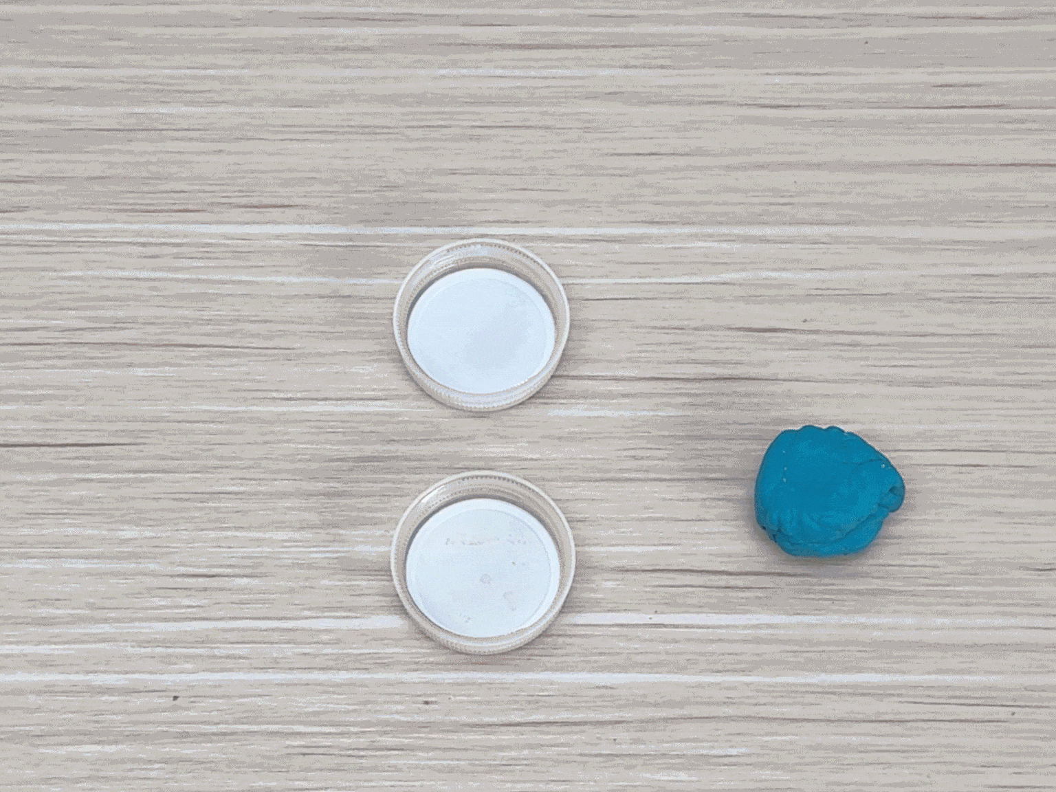 On a light wood-grained surface, two small, white plastic lids or caps are positioned one above the other. To the right of the lids, a small lump of teal-colored putty or modeling clay rests. The simple arrangement suggests these items are ready to be used in a craft or other activity.