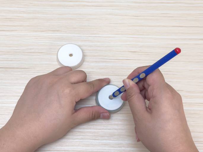 Two hands manipulate small, white plastic discs on a light wood-grained surface. One disc already has a small hole in the center, while the other disc is being worked on. One hand holds the disc steady, while the other hand uses a blue pencil to either enlarge or create a hole in the center. This suggests a stage in a craft project where these discs are being prepared for assembly.