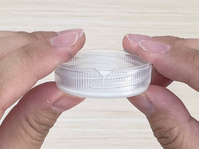 Two hands hold a small, clear plastic lid or container. The lid has a notched or cut-out section on one side and ribbed or textured inner surface. The close-up view emphasizes the detail of the plastic and the way it is being held, likely demonstrating a step in a craft or DIY project.