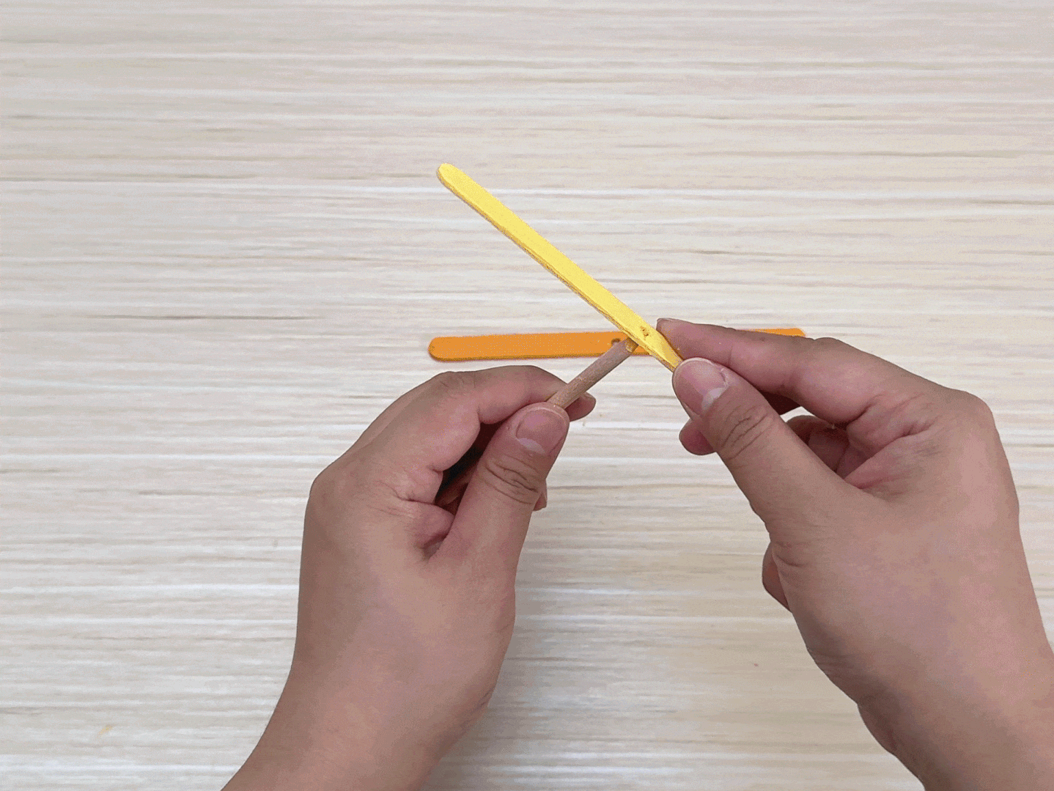 Two hands assemble a small structure made of colorful craft sticks on a light wood grain surface. One hand holds a small, round wooden dowel or stick, while the other hand positions a yellow craft stick with pre-cut slits. An orange craft stick lies nearby. The image captures the process of fitting the yellow stick onto the dowel, suggesting a step-by-step craft project.