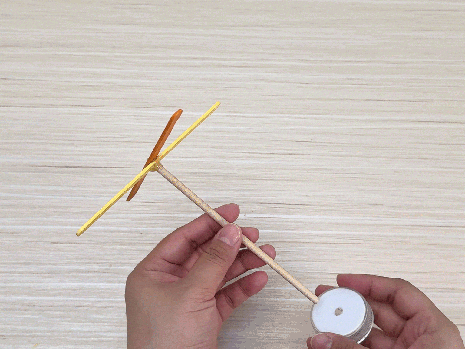 Two hands hold a simple, assembled craft against a light wood grain background. The craft resembles a miniature airplane or windmill. It consists of a central wooden dowel or stick, with two crossed craft sticks – one yellow and one orange – near the top. At the bottom of the dowel, a small white plastic lid or base is attached. The image showcases the completed craft, likely the result of a step-by-step DIY project.