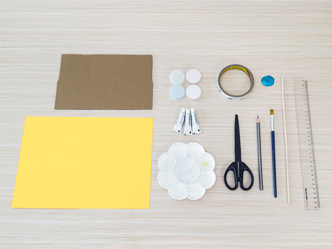 An assortment of craft supplies is laid out on a light woodgrain table. These include a piece of brown cardboard, a sheet of bright yellow paper, three small containers of paint (light blue, light pink, and white), a roll of clear tape, a small blob of teal putty or clay, a thin wooden dowel, a clear plastic ruler, a pencil, a paintbrush, a pair of scissors, three small tubes of glue, and a flower-shaped paint palette with a dab of yellow paint in one section. The items are arranged neatly, suggesting preparation for a craft project.