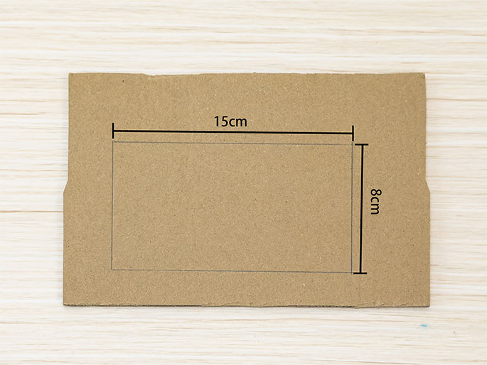 A piece of brown cardboard rests on a light woodgrain table. A rectangular area is marked on the cardboard with thin lines. Labels indicate the dimensions of the marked area: 15cm along the longer horizontal side and 8cm along the shorter vertical side. This suggests a section to be cut or folded.
