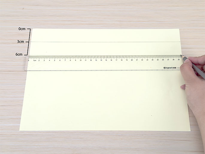 On a light wooden surface, a hand uses a pencil and a clear ruler to draw a line on a pale yellow piece of paper. The ruler is positioned horizontally near the bottom of the paper. Lines are already drawn at 3cm and 6cm from the top edge, as indicated by markings to the left of the paper. The hand is in the process of drawing another horizontal line along the ruler.