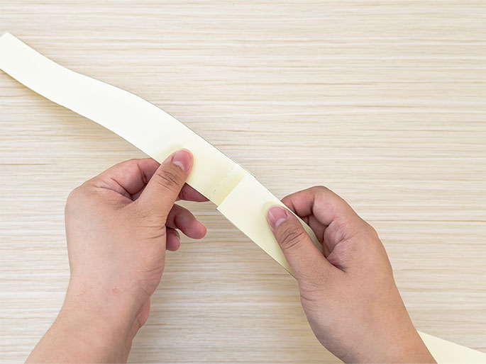 Two hands hold a long, thin strip of pale yellow material over a light woodgrain surface. The strip is slightly curved. A small piece of double-sided tape is visible in the center of the strip, and the hands appear to be in the process of folding the strip at the point where the tape is located.